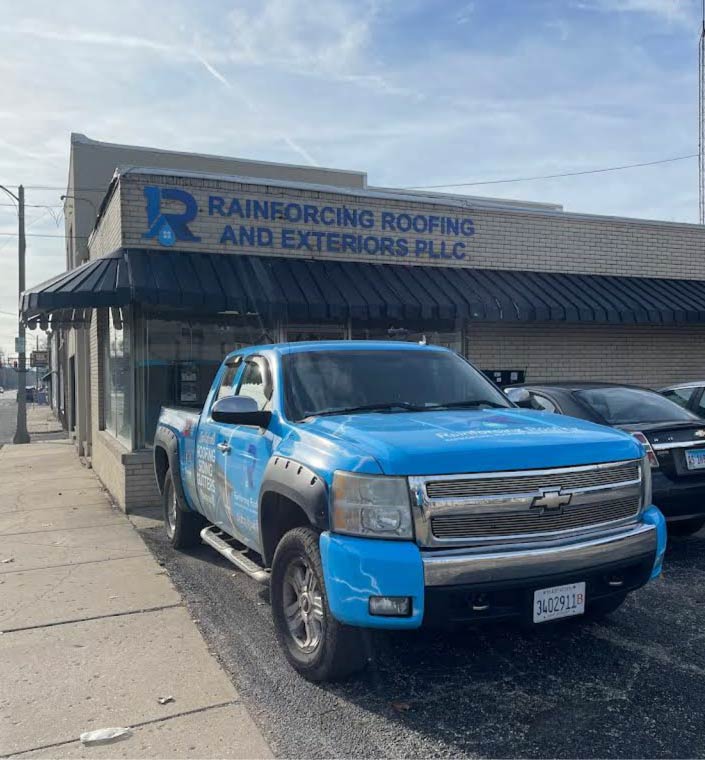 A blue Rainforcing Roofing and Exteriors PLLC branded Chevrolet pickup truck parked in front of the company's brick office building. The truck features a full vinyl wrap with the company logo and service list.