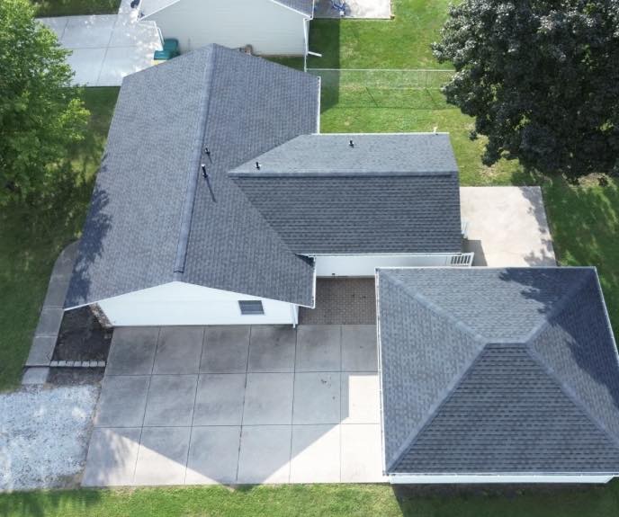 An aerial drone view of a completed residential roofing project in a suburban neighborhood featuring dark gray architectural shingles and clean roof lines.
