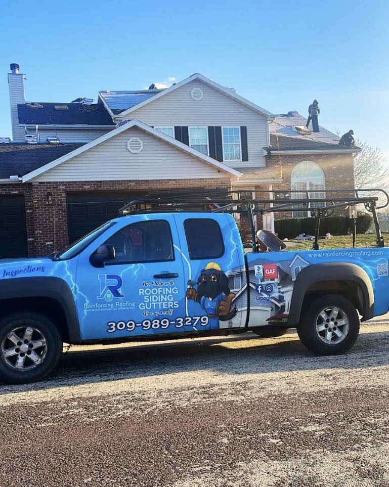 A branded company service truck with a ladder rack and "Rainforcing Roofing" graphics parked at a residential job site. The truck prominently displays the company phone number (309) 989-3279 and GAF certification logo while a crew works on the roof in the background.