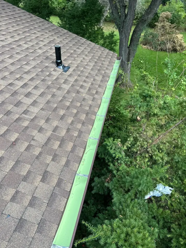 New brown seamless gutter with hangers along a taupe shingle roof with a plumbing vent pipe, trees and lawn below