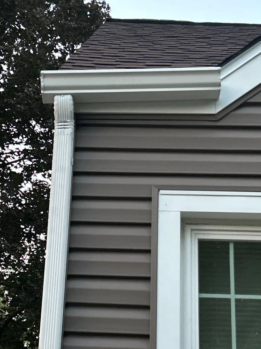 Close-up of a white seamless gutter corner and fluted downspout against brown vinyl siding and white window trim