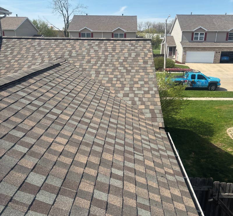 Newly installed blended brown and gray asphalt shingle roof on a residential home with a Rainforcing Roofing truck parked on the street below